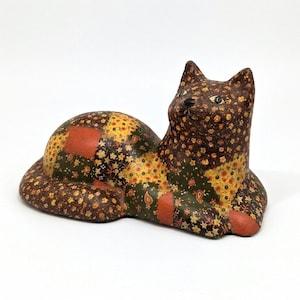 May include: A decorative cat figurine in a resting pose. The cat is covered in a patchwork pattern of brown, yellow, green, and orange floral fabric. The cat's eyes are painted white with black pupils.