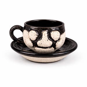 May include: A ceramic teacup and saucer set. The cup and saucer are black and white with a textured, face-like design on the cup. The cup has a rounded handle and sits on a matching saucer. The set is designed for serving tea or coffee.