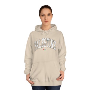 May include: A beige hoodie with a white arch design that says "PALESTINE" and a small Palestinian flag below the text.