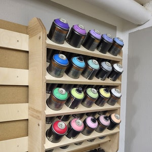 Könnte beinhalten: Ein hölzerner Sprühdosen-Organizer mit mehreren Regalen, der verschiedene Dosen mit farbigen Deckeln enthält. Die Dosen sind schwarz mit unterschiedlich farbigen Deckeln, darunter Lila, Blau, Grün, Gelb und Rosa. Der Organizer ist an einer Wand montiert.