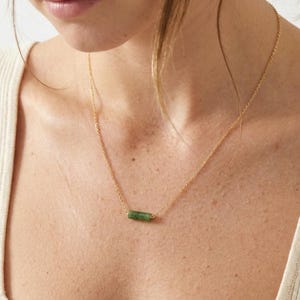 May include: A delicate gold chain necklace featuring a single, small, cylindrical green stone pendant. The necklace rests against the collarbone, showcasing a thin metallic link chain that connects to the horizontal, polished green gemstone bead.