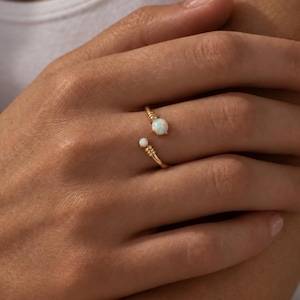 May include: An open-style gold ring featuring two round opal stones of different sizes. The band has a textured, wire-wrapped design at the ends where the opals are set, creating a delicate and balanced look on the finger with a polished finish.