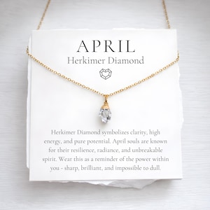 May include: A gold-colored chain necklace with a Herkimer diamond pendant. The pendant is a clear, multifaceted crystal. The necklace hangs from a white card with the word "APRIL" and text about Herkimer diamonds.
