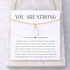 May include: A gold-colored necklace with a teardrop-shaped, white gemstone pendant. The necklace hangs on a white card with the text "YOU ARE STRONG" and inspirational text. The card rests on a light gray surface.