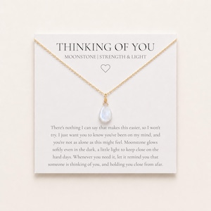 May include: A gold chain necklace with a teardrop-shaped moonstone pendant on a white display card. The card features the text "THINKING OF YOU," "MOONSTONE | STRENGTH & LIGHT," a heart icon, and a printed message about support and connection.