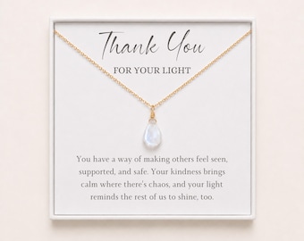 Puede incluir: Un collar de cadena dorada con un colgante de piedra iridiscente en forma de lágrima sobre una tarjeta blanca. La tarjeta presenta el texto Thank You FOR YOUR LIGHT y un mensaje sobre la bondad, el apoyo y la luz en fuentes negras.