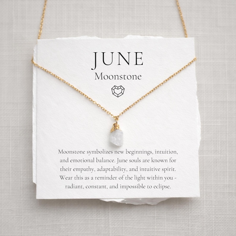 May include: A gold-colored chain necklace with a small, raw moonstone pendant. The necklace hangs in front of a white card that reads "JUNE Moonstone". The card also includes text about moonstone symbolism.