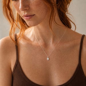 May include: A delicate silver necklace featuring a small, oval-shaped white opal pendant. The pendant is suspended from a thin, polished silver chain that rests against the collarbone, showcasing a smooth, iridescent gemstone setting.