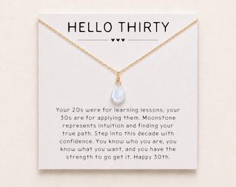 May include: A gold-tone chain necklace with a teardrop-shaped moonstone pendant, displayed on a white card printed with the text HELLO THIRTY and a message about turning thirty, featuring three small black hearts below the title.