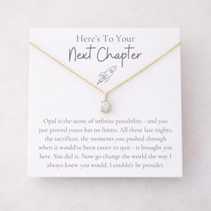 May include: A gold-colored necklace with a small, rectangular opal pendant. The necklace is displayed on a white card with the text "Here's To Your Next Chapter" and a rocket ship illustration. The card also includes inspirational text.