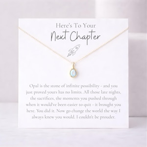 May include: A gold-colored necklace with a small, oval, light blue opal pendant. The necklace is displayed on a white card with the text "Here's To Your Next Chapter" and other inspirational text.