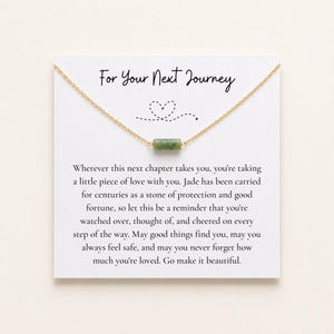 May include: A gold-tone chain necklace featuring a small, cylindrical green jade bead pendant. The necklace is displayed on a white card with the text "For Your Next Journey" and a printed message about protection, love, and good fortune.