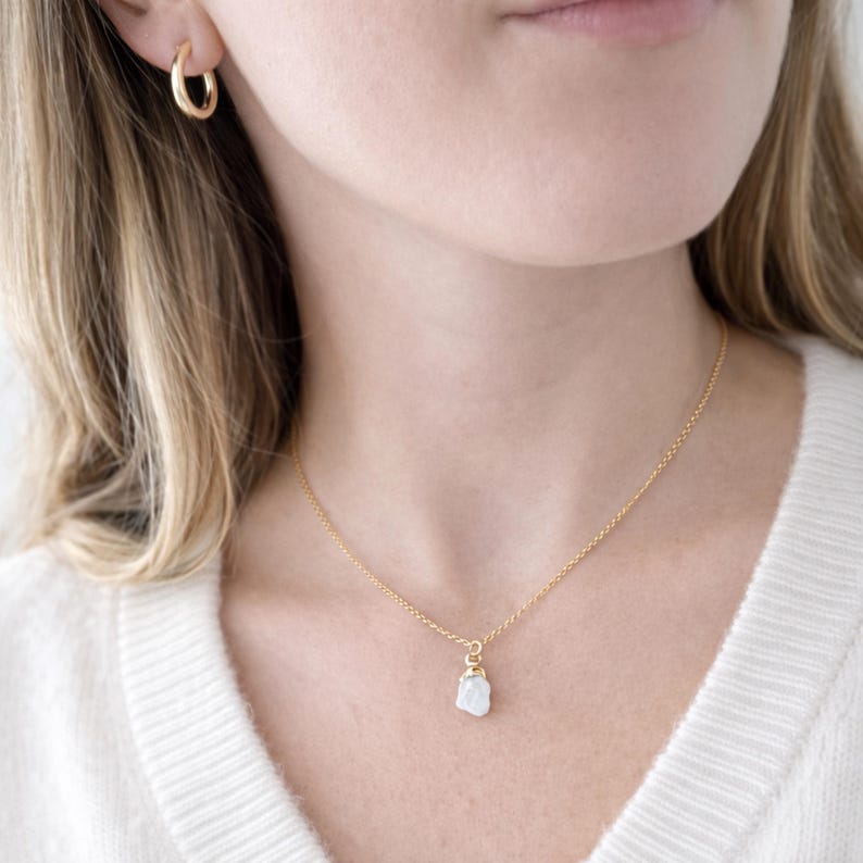 May include: A gold-colored necklace with a small, irregular white stone pendant. The necklace is worn around the neck. A gold hoop earring is visible in the ear. The person is wearing a white sweater.