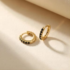 May include: Two gold-colored hoop earrings with black gemstones. The earrings are small and round, with the gemstones set around the outer edge. The earrings are displayed on a light-colored surface.