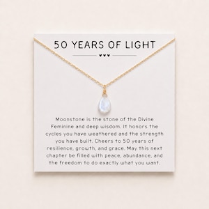 May include: A gold-tone chain necklace with a single teardrop-shaped moonstone pendant, displayed on a white card featuring the text 50 YEARS OF LIGHT and a message about moonstone, resilience, growth, and grace for a fiftieth anniversary.