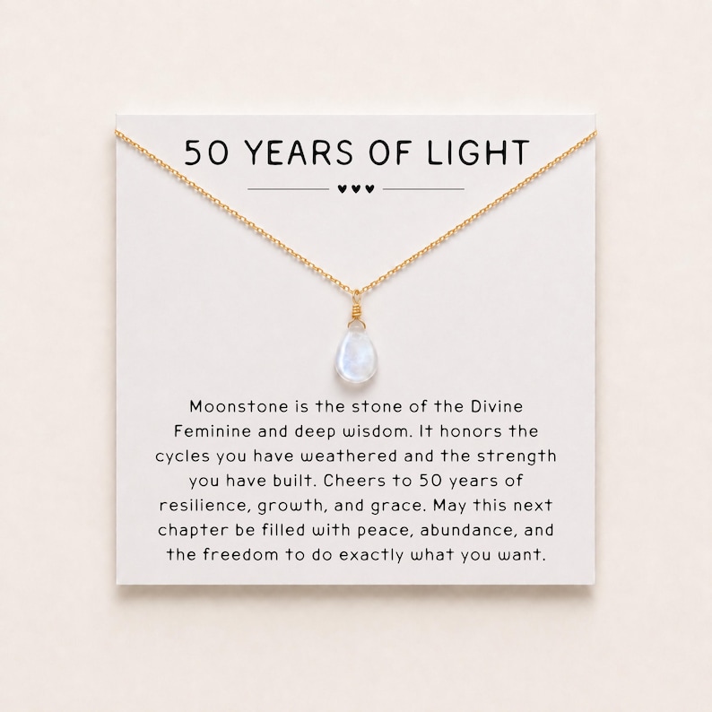 May include: A gold-tone chain necklace with a single teardrop-shaped moonstone pendant, displayed on a white card featuring the text 50 YEARS OF LIGHT and a message about moonstone, resilience, growth, and grace for a fiftieth anniversary.