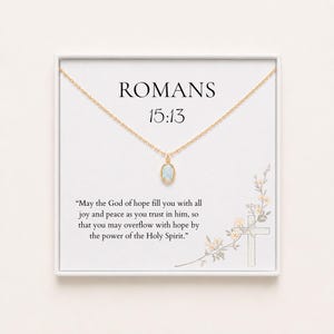 May include: A gold-toned necklace with a small oval pendant featuring a light blue stone. The necklace is displayed on a white card printed with the text ROMANS 15:13 and the verse: May the God of hope fill you with all joy and peace.