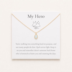 May include: A gold chain necklace with an oval opal pendant displayed on a white card featuring the text My Hero, a mountain graphic, and a message about opal carrying light and someone waiting back home for the wearer to return safely.