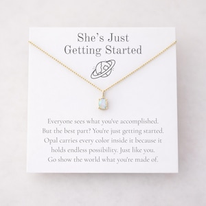 May include: A gold-colored necklace with a small, oval opal pendant. The pendant hangs from a delicate chain. The necklace is displayed on a white card with the text "She's Just Getting Started" and inspirational text.