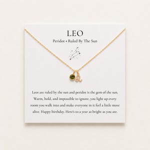May include: A gold-tone chain necklace featuring a small green peridot gemstone charm and a Leo zodiac symbol charm. The necklace is displayed on a white card printed with the text LEO, Peridot, Ruled By The Sun, and a birthday message.