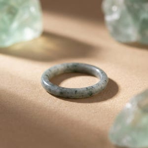 May include: A jade ring with a speckled pattern in shades of green and gray. The ring is set against a neutral background, with soft lighting and a few green stones in the background.