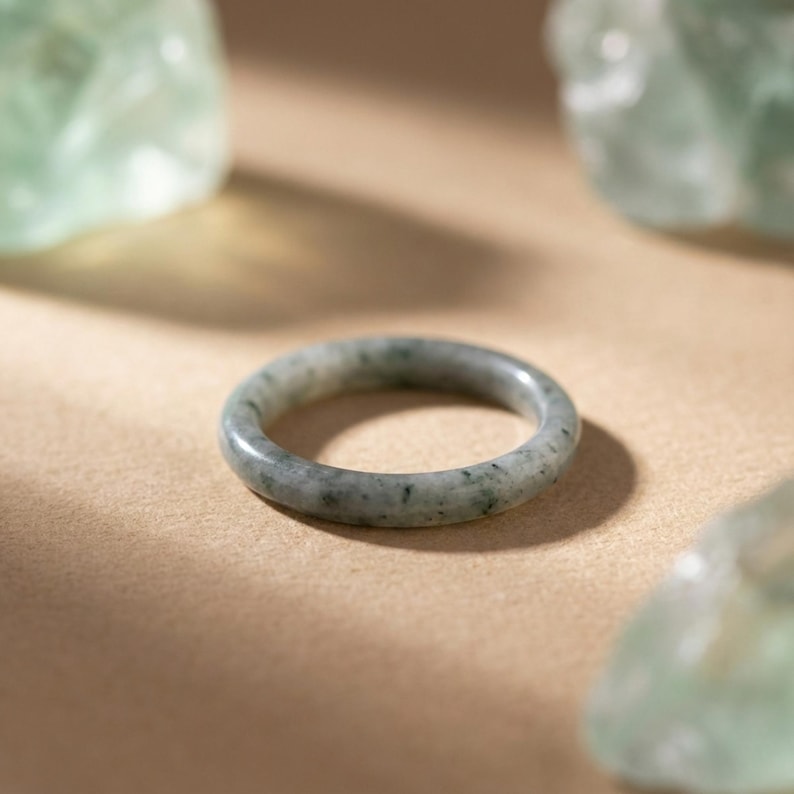 May include: A jade ring with a speckled pattern in shades of green and gray. The ring is set against a neutral background, with soft lighting and a few green stones in the background.