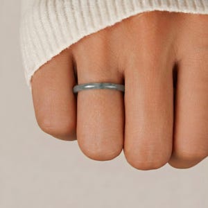 May include: A close-up of a light green jade ring. The ring is a simple band design, showcasing a smooth, polished surface. The ring is worn on a finger, with a soft, neutral background.