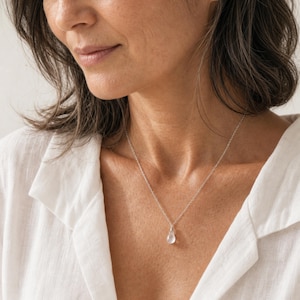 May include: A delicate silver chain necklace featuring a small, translucent, teardrop-shaped pendant. The pendant is clear with a smooth, polished surface, suspended from a thin, metallic silver link chain that rests against the collarbone area.