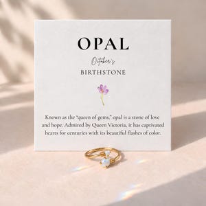 May include: A gold-toned wire ring featuring two small, iridescent opal stones, displayed in front of a white card printed with the text OPAL October's BIRTHSTONE, a small purple flower illustration, and a paragraph about the gemstone.