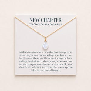 Puede incluir: Un collar de cadena dorada con un colgante de piedra lunar en forma de lágrima. El collar se presenta sobre una tarjeta blanca con el texto NEW CHAPTER The Stone for New Beginnings y un párrafo sobre los ciclos y cambios de la vida.