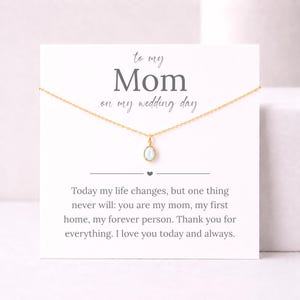 May include: A gold-toned chain necklace with a small, oval-shaped iridescent pendant displayed on a white card. The card features the text: to my Mom on my wedding day. Today my life changes, but one thing never will: you are my mom, my first home.