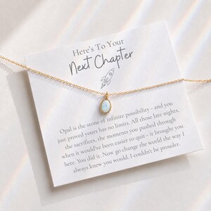 May include: A gold-tone chain necklace with a small, oval-shaped opal pendant. The necklace is displayed on a white card featuring the text 'Here's To Your Next Chapter' and a rocket ship icon, along with a printed message about opal stones.