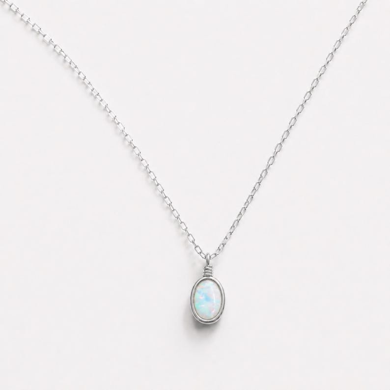 May include: A silver-toned necklace featuring a delicate chain and a single oval-shaped pendant. The pendant contains a smooth, iridescent white stone with subtle blue and pink flashes, secured by a simple wire-wrapped metal setting at the top.