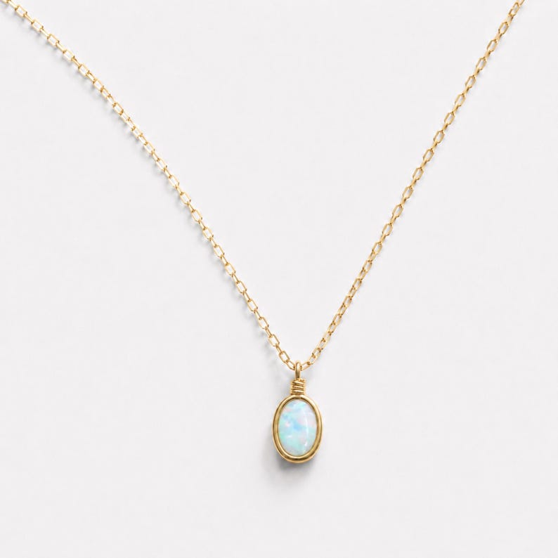 May include: A delicate gold chain necklace featuring a small, oval-shaped opal pendant. The pendant is secured in a simple gold wire setting with a wrapped bail at the top, showcasing the iridescent white and blue tones of the gemstone.