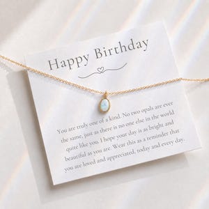 May include: A gold-tone chain necklace with a small, oval-shaped iridescent opal pendant. The necklace is displayed on a white card featuring the text Happy Birthday and a printed message about being one of a kind, bright, and beautiful.