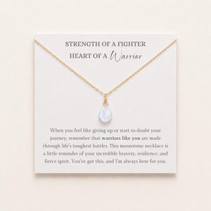 Pode incluir: Um colar de corrente dourada com um pingente de pedra da lua em forma de gota num cartão branco. O cartão apresenta o texto STRENGTH OF A FIGHTER HEART OF A Warrior e um parágrafo sobre bravura, resiliência e um espírito feroz.
