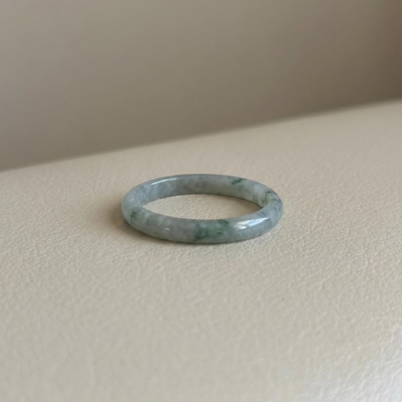 May include: A jade ring with a smooth, polished surface. The ring is a pale green color with darker green marbling throughout. The ring is a simple band design.