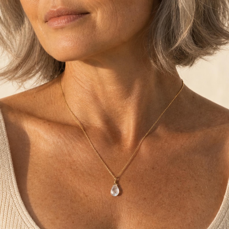 May include: A delicate gold chain necklace featuring a single, clear, teardrop-shaped pendant. The pendant hangs centrally from the thin, polished gold-tone metal chain, which rests against the collarbone area, creating a simple and refined look.
