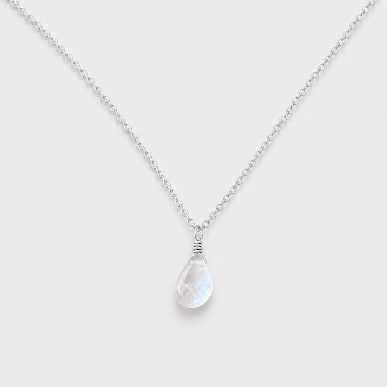 May include: A silver chain necklace featuring a single teardrop-shaped moonstone pendant. The translucent, iridescent stone is secured with a silver wire wrap at the top, connecting it to a delicate, polished silver link chain.