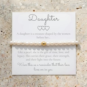 May include: A white pearl bracelet with a gold bead is displayed on a white card. The card reads "Daughter" with a message about a daughter's legacy. The background is a light beige, textured surface.