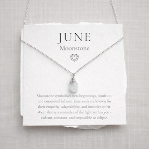 May include: A silver necklace with a small, raw moonstone pendant hangs in front of a white card. The card reads "JUNE Moonstone" in black text. The necklace is displayed on a white background.