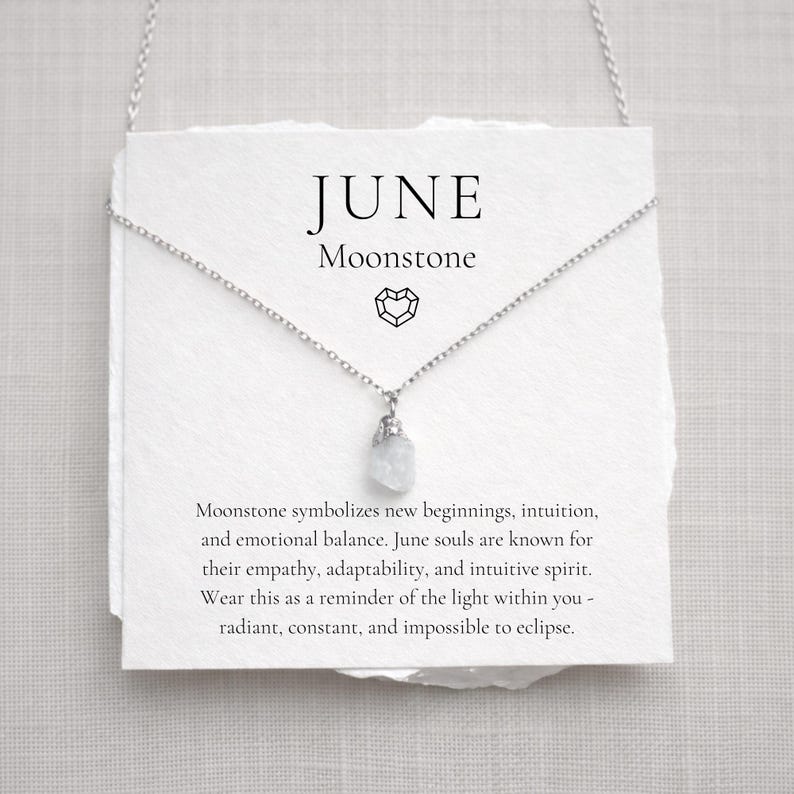 May include: A silver necklace with a small, raw moonstone pendant hangs in front of a white card. The card reads "JUNE Moonstone" in black text. The necklace is displayed on a white background.