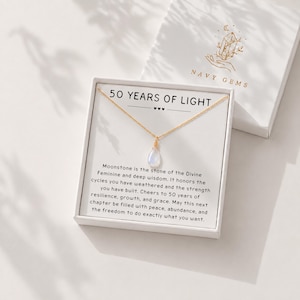 May include: A gold chain necklace with a teardrop-shaped moonstone pendant presented in a white gift box. The box insert features the text 50 YEARS OF LIGHT and a message about moonstone, with the brand name NAVY GEMS printed on the lid.