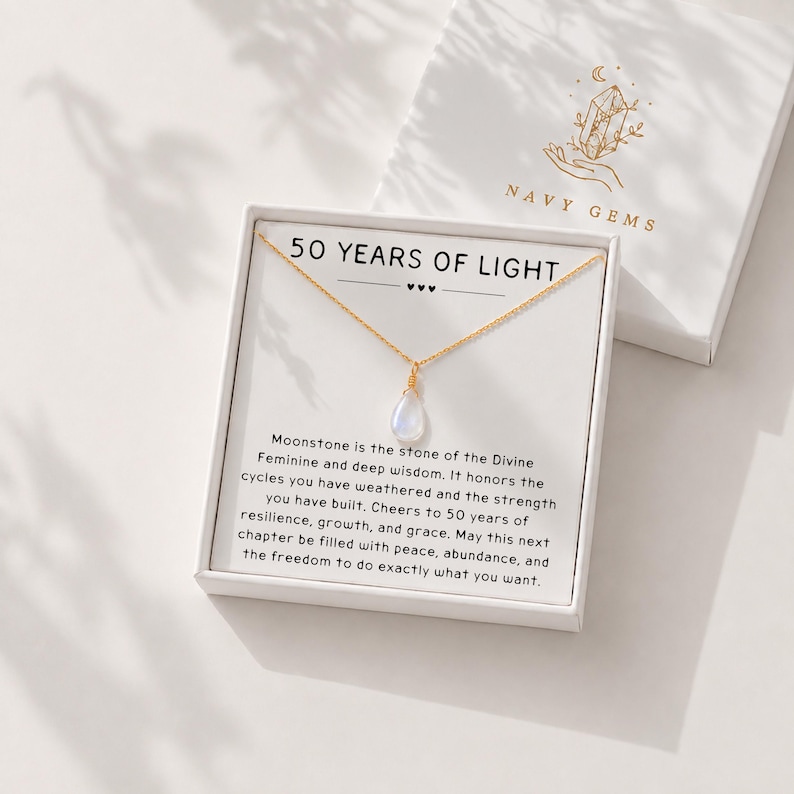 May include: A gold chain necklace with a teardrop-shaped moonstone pendant presented in a white gift box. The box insert features the text 50 YEARS OF LIGHT and a message about moonstone, with the brand name NAVY GEMS printed on the lid.