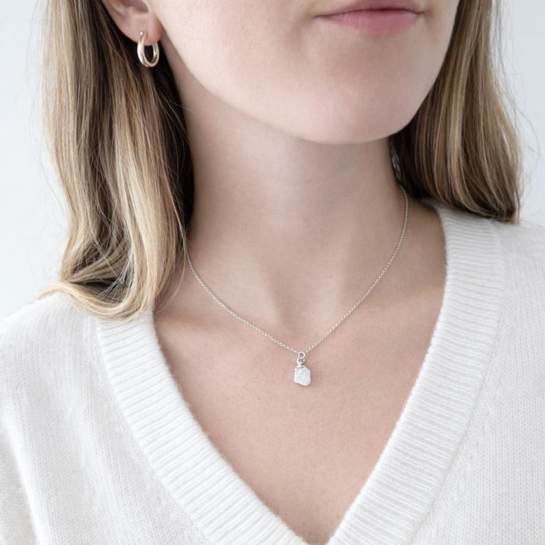 May include: A silver necklace with a small, irregularly shaped pendant. The pendant appears to be a light-colored stone. The model is wearing a white sweater and gold hoop earrings. The necklace is centered on the chest.