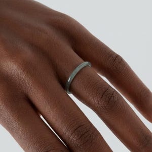 May include: A slender, light green ring is worn on a finger. The ring has a smooth, rounded band and a subtle, natural color. The ring's design is simple and elegant, suitable for everyday wear.