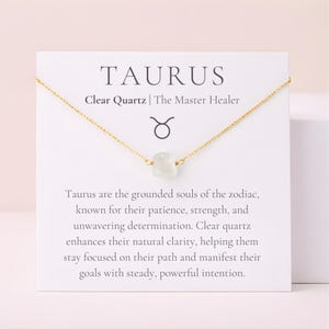 May include: A gold-colored chain necklace with a clear quartz pendant. The necklace is displayed on a white card with the word "TAURUS" and the text "Clear Quartz | The Master Healer". The card also features the Taurus zodiac symbol.