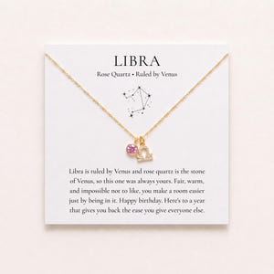 May include: A gold-tone chain necklace featuring a pink circular gemstone charm and a small Libra zodiac symbol pendant. The necklace is displayed on a white card printed with the text LIBRA, Rose Quartz, and a birthday message.