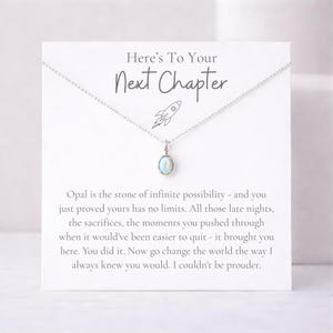 May include: A silver necklace with an oval opal pendant, displayed on a white card. The card reads "Here's To Your Next Chapter" with a rocket ship illustration and inspirational text about new beginnings.