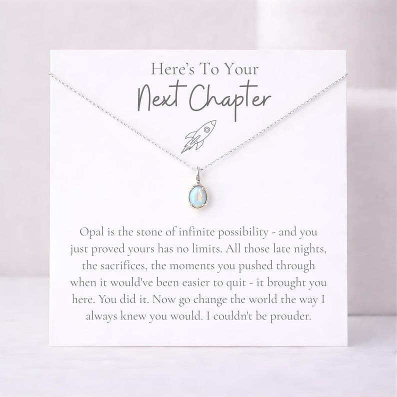 May include: A silver necklace with an oval opal pendant, displayed on a white card. The card reads "Here's To Your Next Chapter" with a rocket ship illustration and inspirational text about new beginnings.